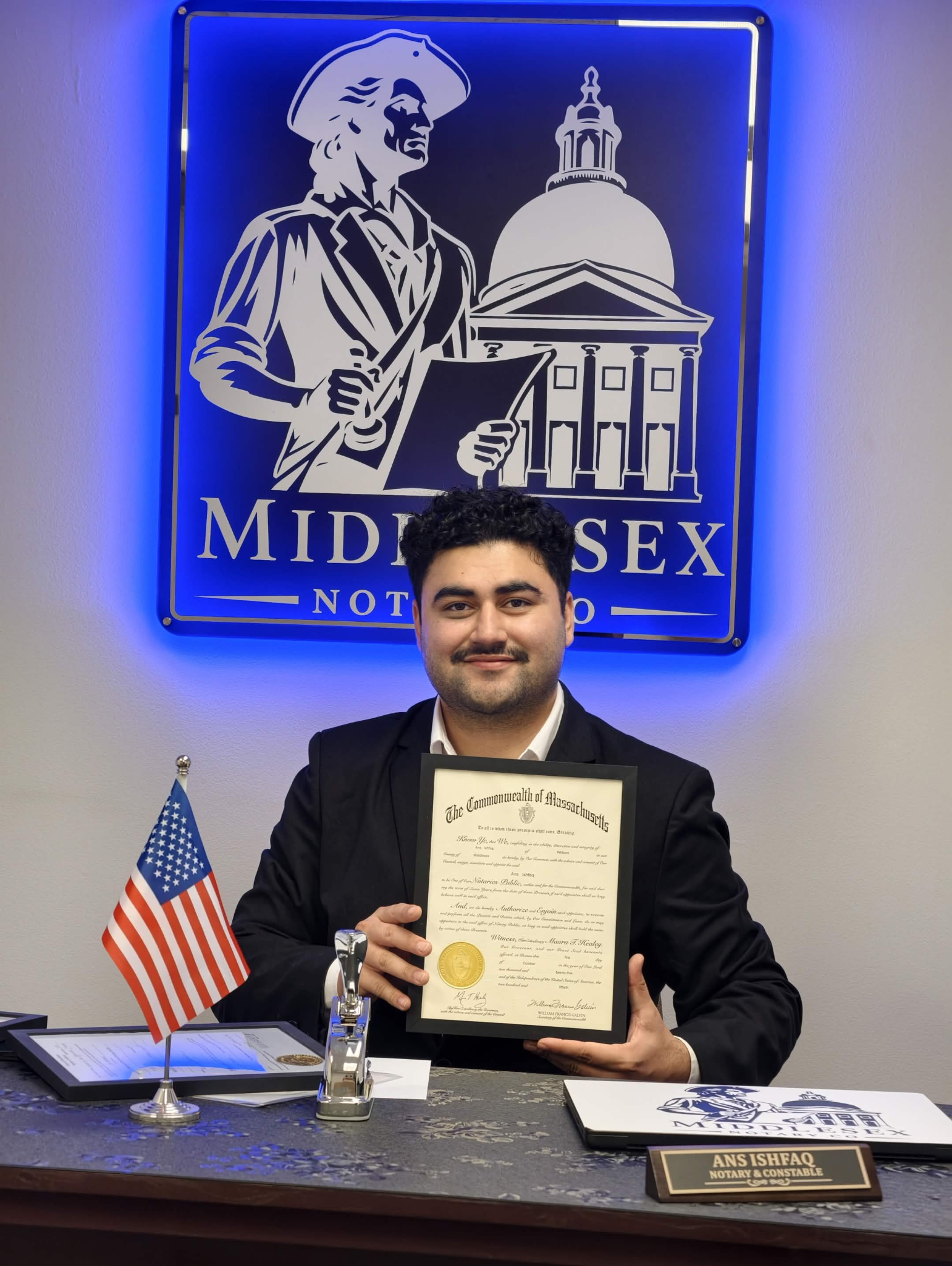Ans Ishfaq smiling at his Middlesex Notary office while holding his Massachusetts notary commission.